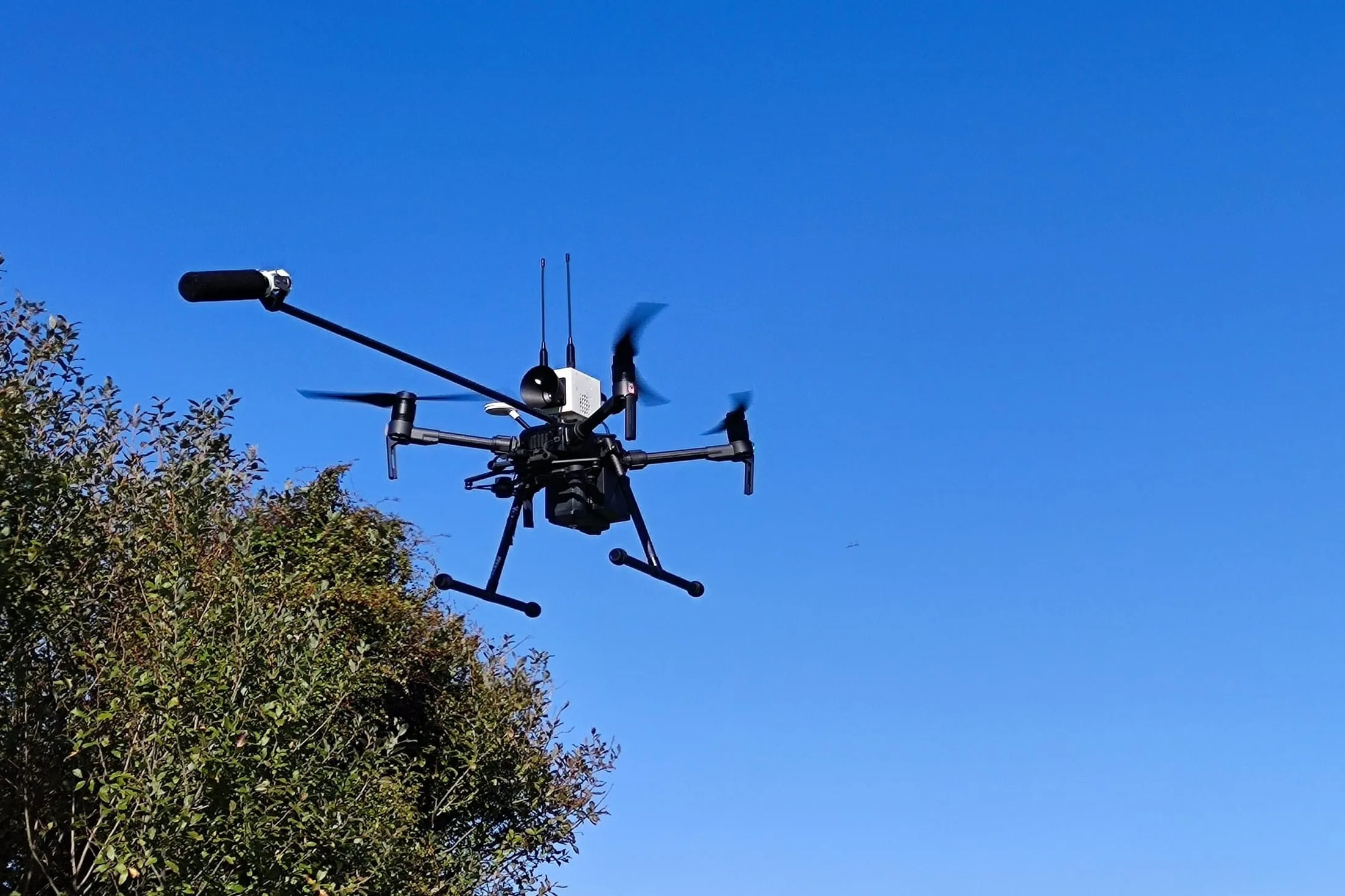 DJI M210 in flight against clear sky, carrying the array payload with a microphone on a forward boom and mesh radio antennas.